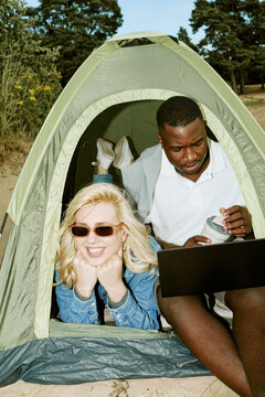 Vertical shot of young adult Caucasian woman and African American man relaxing in camping tent with laptop. Digital nomad lifestyle concept for travel advertising