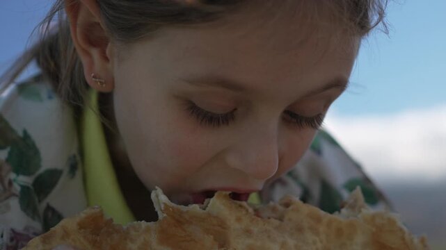 Child eating food, Young girl enjoying large traditional fried pastry outdoors under a clear blue sky.