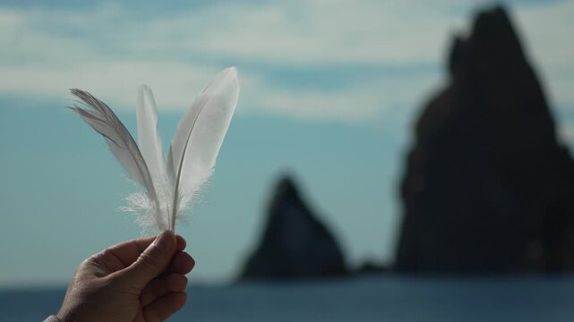 Feathers hand nature held gently against dark sea stacks and peaceful blue sky symbolizing lightness.