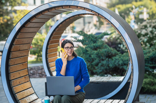 Businesswoman talks on smartphone while working on laptop outdoors