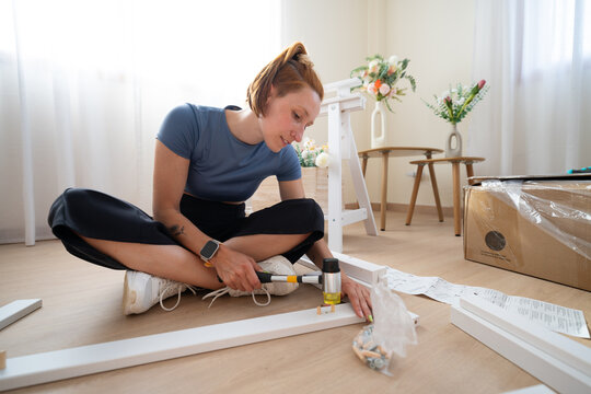 A woman sits on the floor assembling new furniture with a hammer