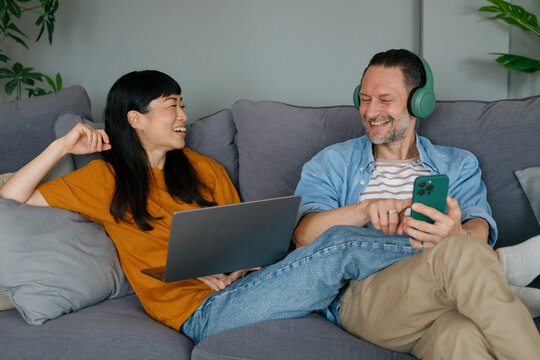 Couple sitting on sofa using laptop and smartphone, smiling and relaxing