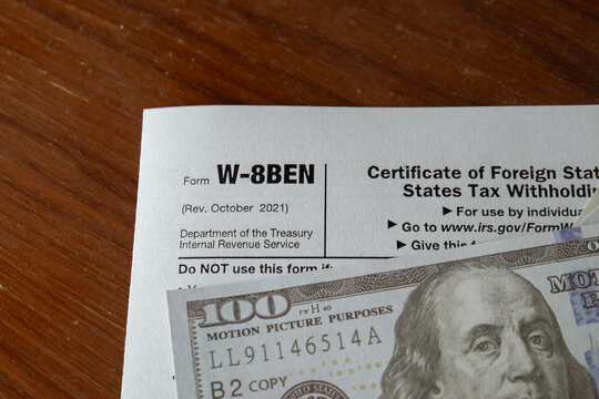 Close up of US IRS Form W-8BEN for foreign status tax withholding with US dollar prop money on a wooden desk. Concept of international tax, foreign income, freelance payment, and accounting.