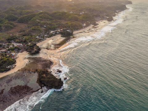 Aerial view of Gunungkidul Regency coastline with rocky cliffs, sandy beaches, and waves crashing against the shore under golden hour light in Special Region of Yogyakarta, Indonesia.