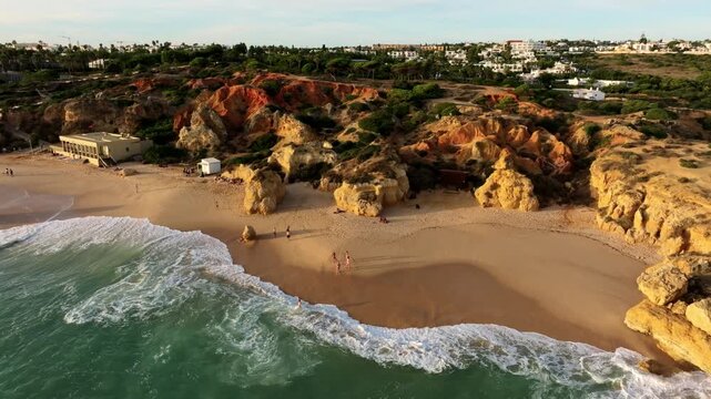 Drone view of Praia de Sao Rafael beach in Algarve, Portugal, with foamy surf, sand, golden cliffs, and a beach building. Orbit parallax shot