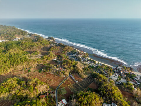 Aerial view of Gunungkidul Regency coastline featuring lush green hills, agricultural fields, and a winding road leading to the ocean, Special Region of Yogyakarta, Indonesia.