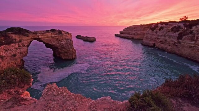 Dramatic sunset view of Arco de Albandeira at Praia de Albandeira in Algarve, Portugal, with pink sky, limestone sea arch, Atlantic water, and low tide foam patterns near the rocks