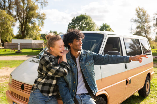 A young couple leans against a camper van and points at nature