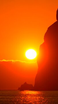 Sunset sea cliff silhouette against a vibrant orange sky over calm ocean waves at dusk during a peaceful evening