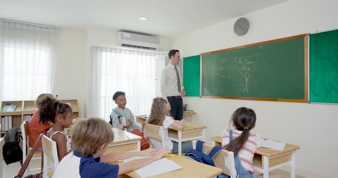 Wide elementary classroom scene follows male teacher guiding chalkboard lesson while young pupils sit at wooden desks, write in notebooks, raise pencils and settle into back to school learning routine