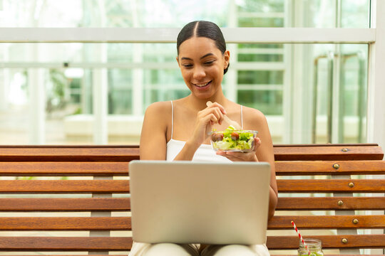 Smiling woman eating salad and working on laptop outdoors