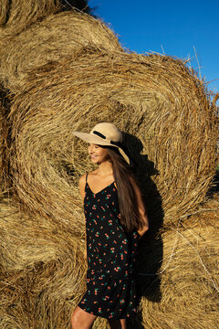 A smiling woman wears a hat near a haystack in a sunny summer field