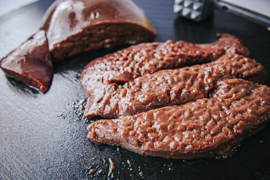 Raw liver pieces arranged on a dark cutting board beside a metal meat tenderizer, showing glossy surfaces and varied organ-meat textures.
