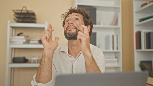 Man with laptop and fingers crossed at a desk in a building office, looking upward amid bookshelves and stacked folders; hope focus.