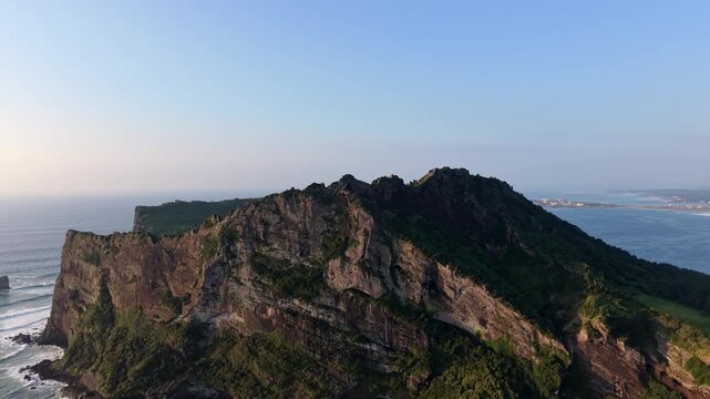 Aerial view of Seongsan Ilchulbong volcanic tuff cone rising above the ocean with rugged cliffs and green vegetation under a clear sky in Seogwipo-si, Jeju-do, South Korea.