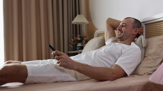 Man lays on bed, switches channels with remote control, watches TV and smiles.