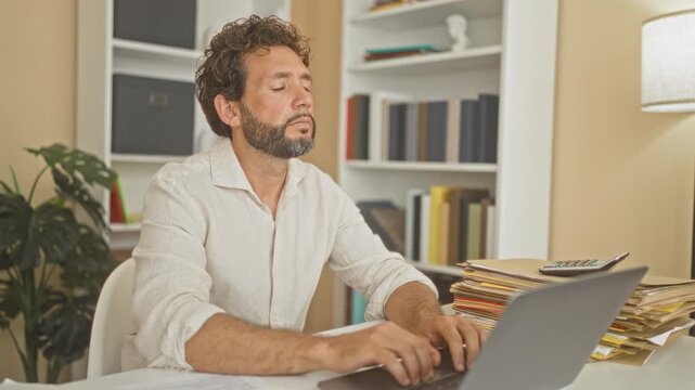Man middle aged at laptop resting head on folded arms at desk surrounded by folders and books in building; work fatigue.