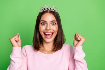 Young woman with tiara celebrates fashion fun with pink sweater and bright smile against green background © deagreez
