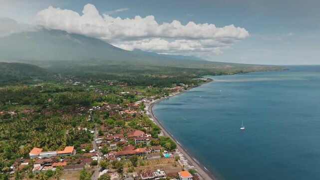 Amed village with its traditional buildings and lush palm trees lining dark sand beach along tropical ocean, towering mount Agung volcano under a cloudy sky. Bali, Indonesia. Aerial view drone flight
