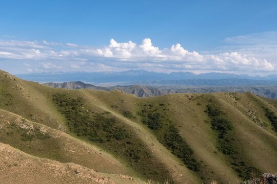 A wide panoramic view of green rolling hills and a distant mountain range under a blue sky with fluffy white clouds. 