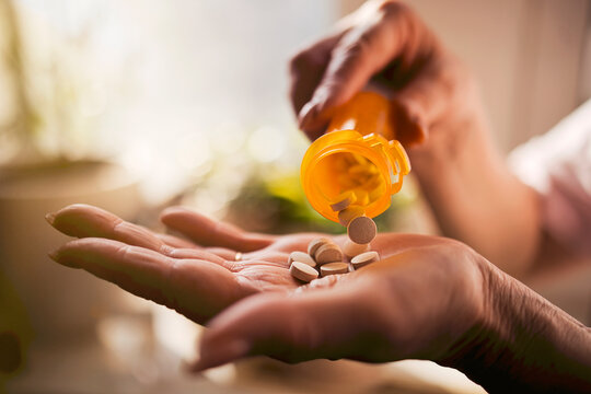 Hand holding a palm with white pills as a yellow prescription bottle tips over, spilling medication in a bright indoor setting with plants in the background