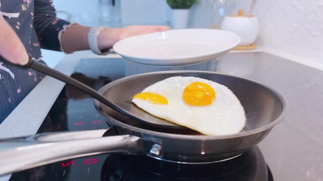 A person uses a black spatula to lift a cooked fried egg from a pan on a stove into a plate. This warm morning routine creates a cozy and pleasant feeling.