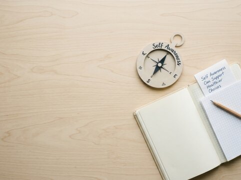 Flat lay top view of a paper compass labeled Self Awareness, an open blank notebook, and a pencil on a light wood surface with a motivational sticky note.