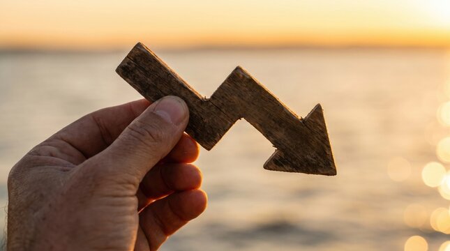 Wooden arrow pointing down held by hand, financial loss and economic decline concept, business failure symbol at sunset