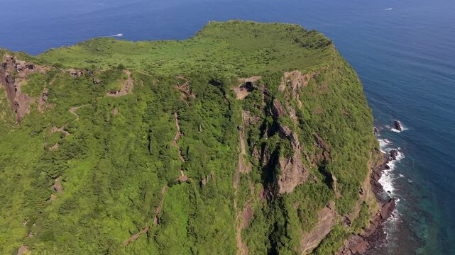 Aerial view of Seongsan Ilchulbong volcanic crater with lush green vegetation, steep cliffs, and surrounding blue ocean waters in Seogwipo, Jeju-do, South Korea.