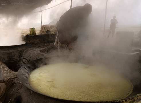 View of a worker stirring boiling sugarcane juice in a large metal vat to produce traditional jaggery in a rustic, smoky outdoor workshop in Multan, Punjab, Pakistan.