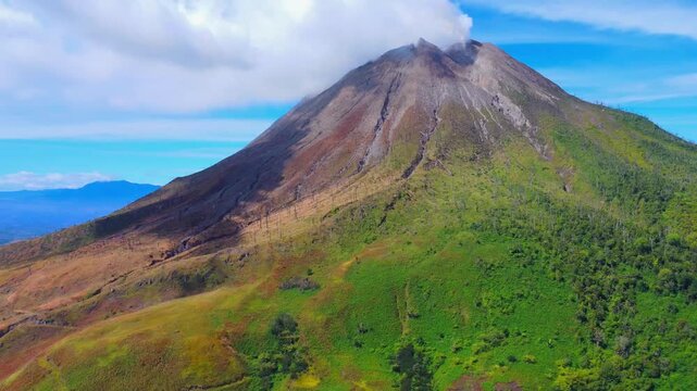 Aerial view of Mount Sinabung volcano with smoke rising from the crater, lush green slopes, and rocky terrain in Naman Teran, North Sumatra, Indonesia.