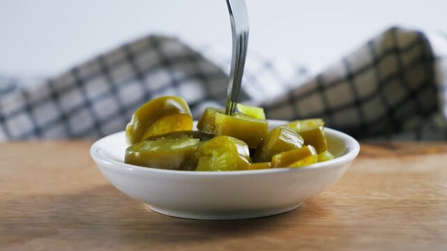 Fork picking up pickled jalapeno slices from a white bowl on wooden table, checkered napkin background, close-up food video footage. High quality 4k footage