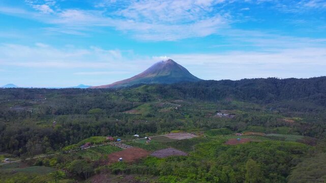Aerial view of Mount Sinabung with its volcanic peak rising above lush green agricultural fields and dense forests under a clear blue sky in Naman Teran, North Sumatra, Indonesia.