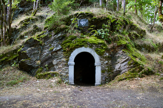 Ein steinernes Portal mit dunklem Eingang liegt in einer moosbedeckten Felswand am Waldrand; eine kleine Tafel ist oberhalb angebracht.