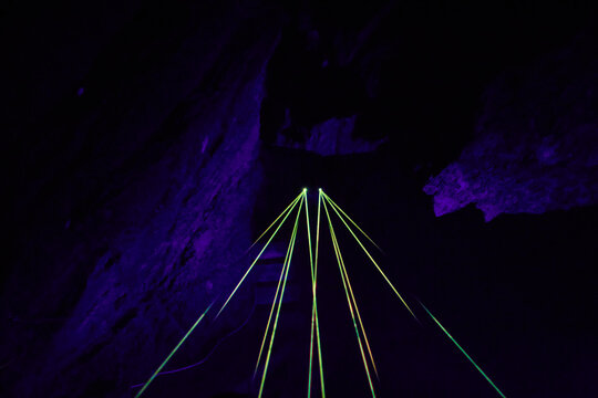 Mehrere gr&uuml;ne und gelbe Laserstrahlen verlaufen perspektivisch durch einen dunklen H&ouml;hlengang der Drachenh&ouml;hle Syrau.