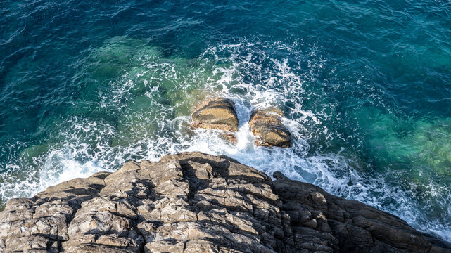 Aerial view of rocky cliffs meeting the deep blue sea with crashing waves and white foam in Italy.