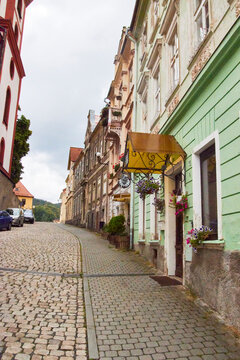 Eine ansteigende Kopfsteinpflasterstra&szlig;e f&uuml;hrt durch die historische Altstadt von Loket; farbige Fassaden, Blumenampeln und geparkte Autos s&auml;umen den Stra&szlig;enraum.