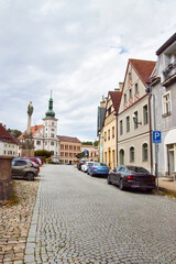 Obraz premium Eine gepflasterte Straße führt durch das historische Zentrum von Loket zu einem Uhrturm und einer barocken Säule; entlang der Häuser stehen Fahrzeuge.