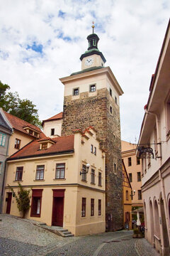 Ein steinerner Uhrturm mit gr&uuml;n patinierter Dachhaube ragt zwischen historischen H&auml;usern und roten Ziegeld&auml;chern der Altstadt von Loket auf.