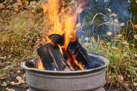 Hohe orangefarbene Flammen brennen zwischen verkohlten Holzscheiten in einer steinernen Feuerschale; im Hintergrund sind Gras und ein Baumstamm sichtbar.