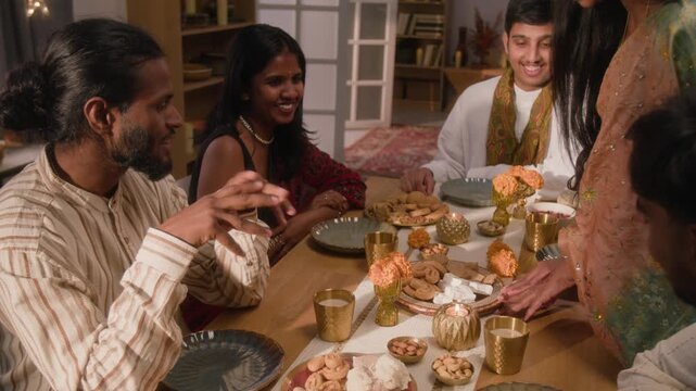 Indian young woman serving traditional sweets to friends at festive dinner table, camera zooming out. Footage suitable for Diwali holiday advertising, cultural campaigns, and food brand marketing
