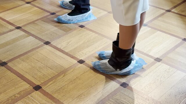 Visitors wearing blue shoe covers walk across a wooden parquet floor inside Catherine Palace in Pushkin, Saint Petersburg. The historical hall fills with a peaceful sense of quiet respect.
