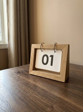 Wooden desk calendar showing the number zero one on a white card, placed on a dark wooden table near a window with beige curtains in the room