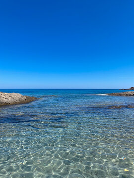 Scenic view of a Mediterranean paradise bay in Corsica, France, featuring transparent crystalline water with sunlight reflections on the sandy bottom and low coastal rocks under a clear blue sky. 