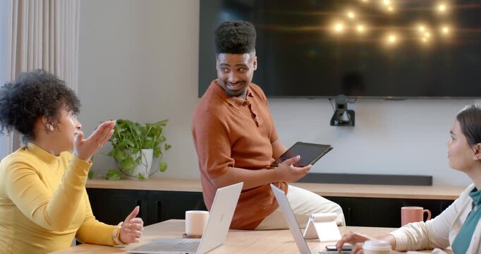 Diverse coworkers watching man showing tablet one woman gesturing other typing on laptop in office