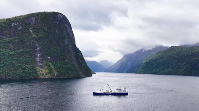 Aerial view of a wellboat navigating through the calm waters of a fjord surrounded by steep mountains under a cloudy sky in Liabygda, More og Romsdal, Norway.