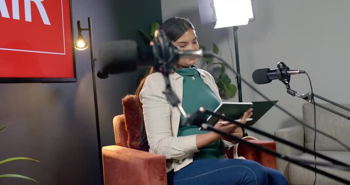 African American woman glancing at mics, opening folder, flipping pages writing notes for podcast