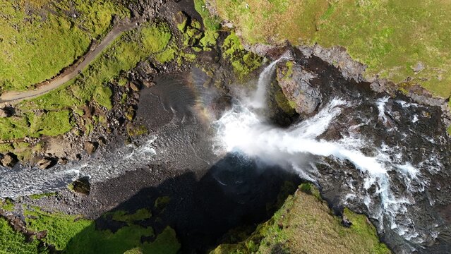 Aerial view of a powerful waterfall cascading over rocky cliffs into a pool with a faint rainbow in the mist, surrounded by vibrant green mossy terrain in Iceland.