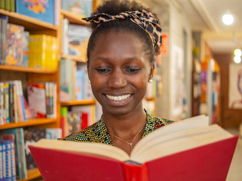 Happy Black female student reading a red book near bookshelves in a library.