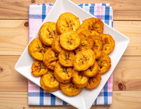 Golden fried plantains tostones on white plate with colorful napkin, Caribbean Latin American snack food, top view on wooden table
